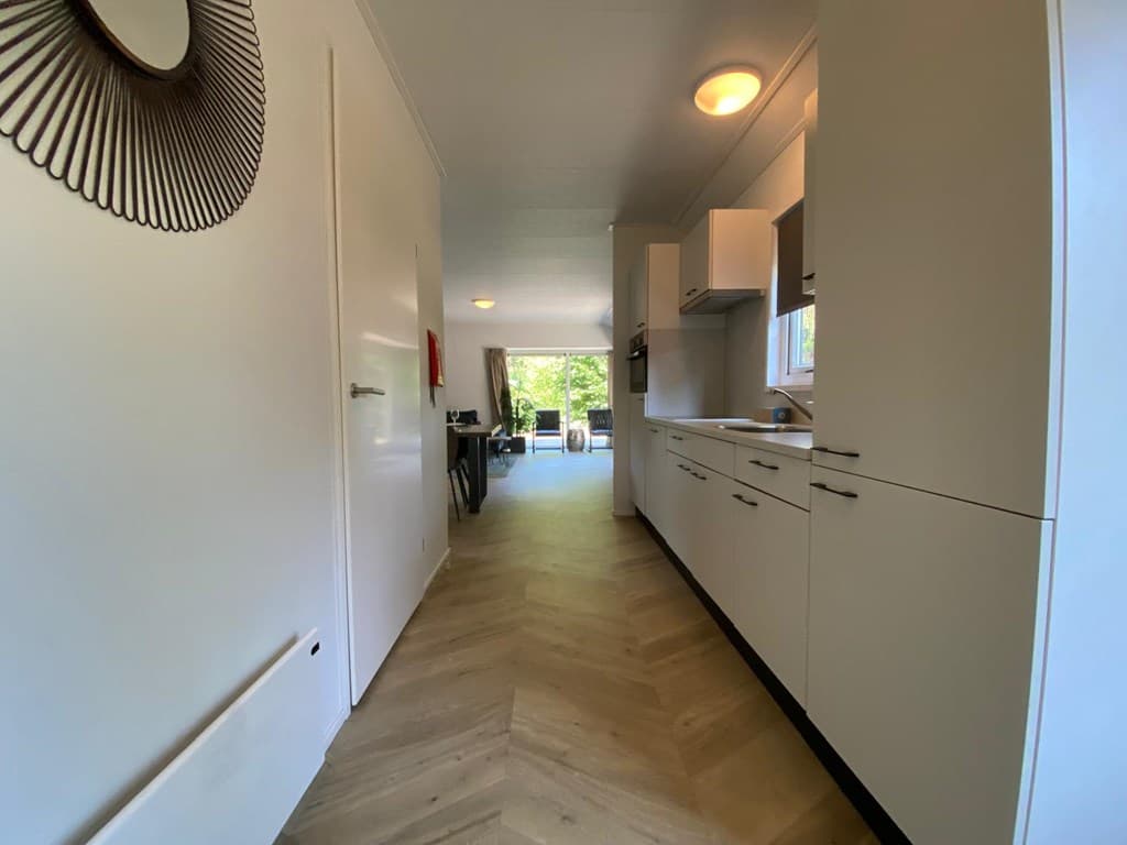 Ingang van de woning met een lange gang, lichte keuken en zicht op een eetgedeelte met grote ramen en veel natuurlijk licht.