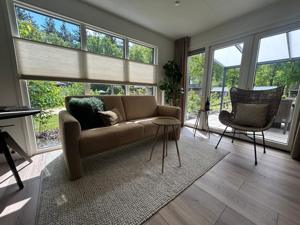 Gezellige woonkamer met bank, vloerkleed, planten en grote ramen met uitzicht op de tuin en het terras.