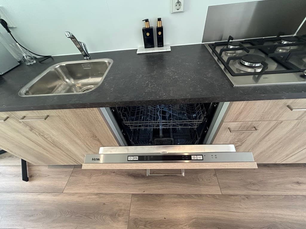 Keuken met een zwart blad, diepe spoelbak, gaskookplaat en open vaatwasser in een houten kastenwand.