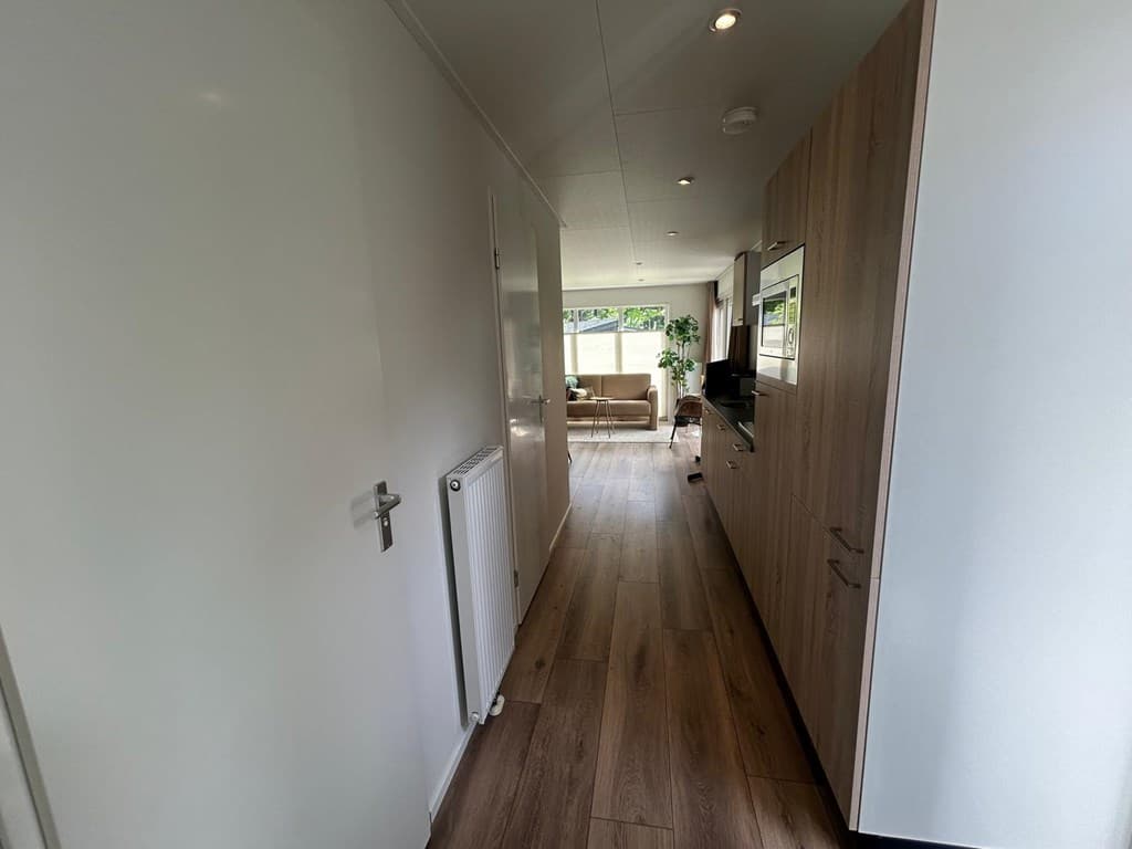 Lange gang met laminaatvloer en lichte muren, aan de rechterzijde een kast en radiator, aan het einde zicht op woonkamer met 