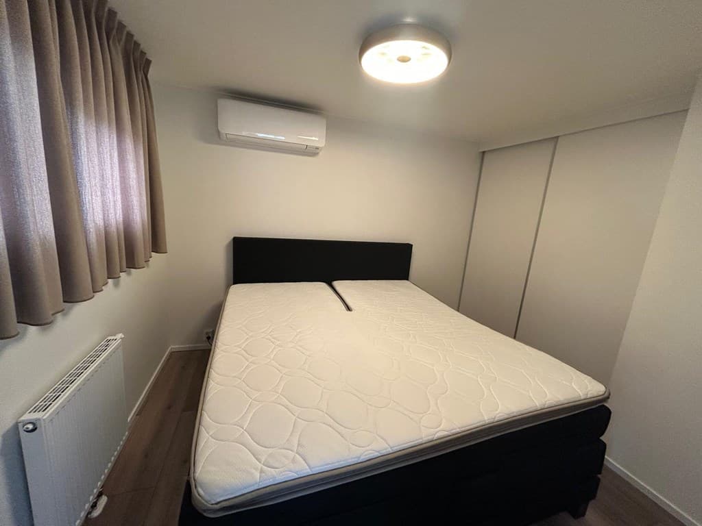 Slaapkamer met zwart bed, airconditioning, radiator en inbouwkast, gordijnen aan het raam.