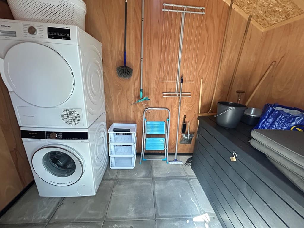 Wasruimte met wasmachine, droger, schoonmaakspullen en een opbergmeubel op een vloer van grijze tegels