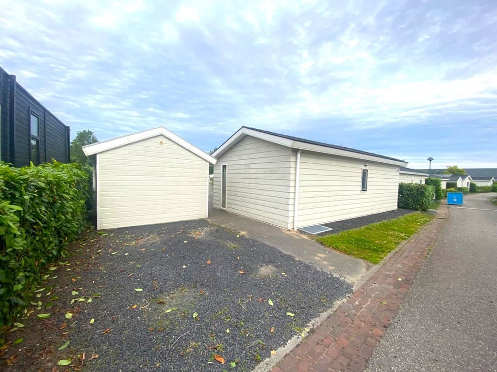 Twee vakantiewoningen met een oprit, tuin en en een schuur, omringd door groene heggen en bestrating.