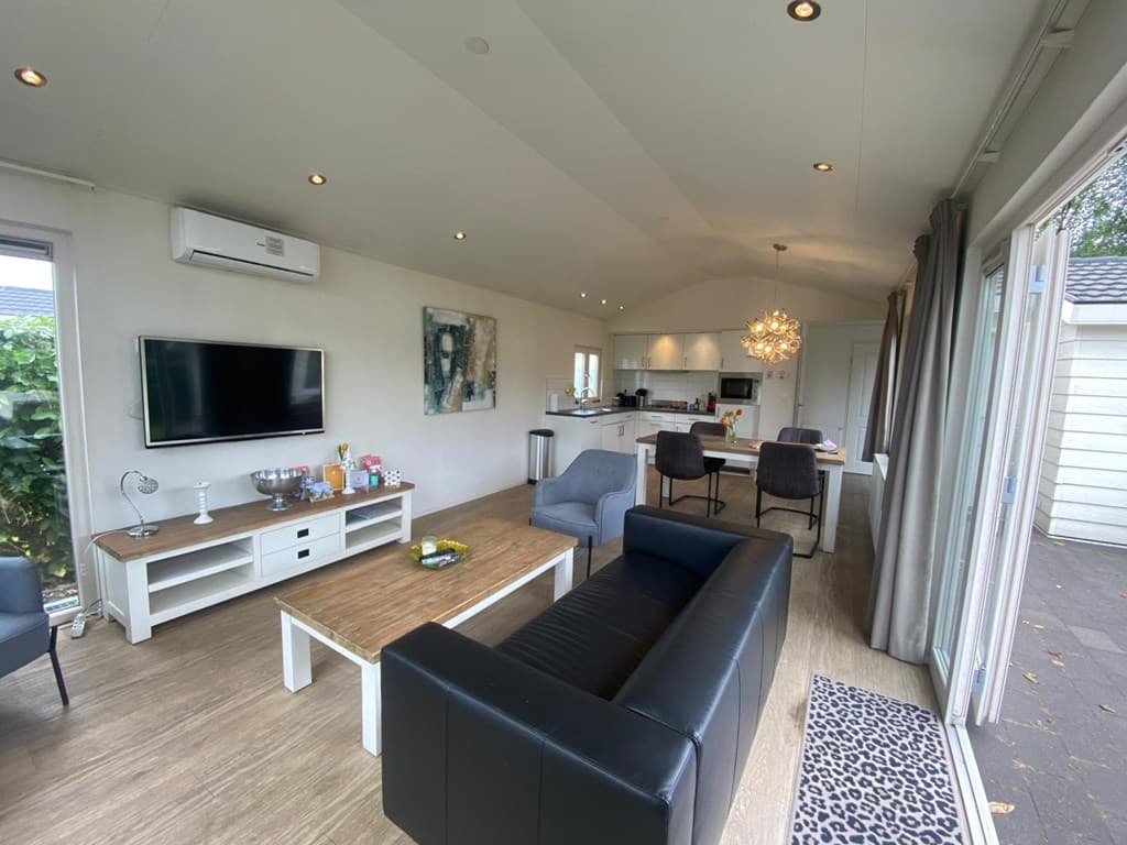 Moderne woonkamer met open keuken, bank en eetruimte, grote ramen met uitzicht op een tuin, lichte inrichting en unieke lamp.