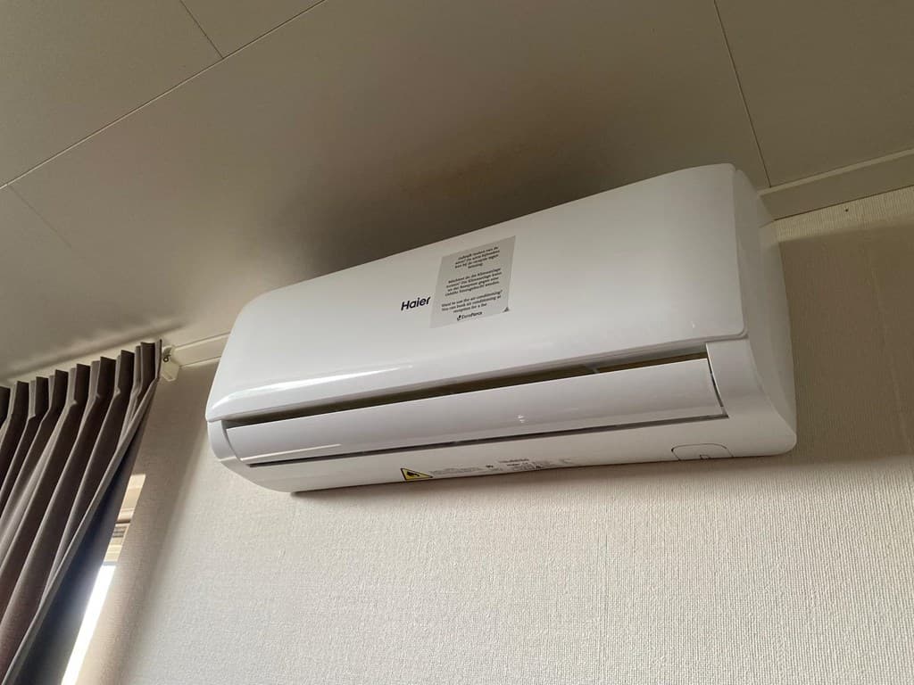 Airconditioningseenheid aan de muur met een handleiding in een kamer met gordijnen en een lichte wand.