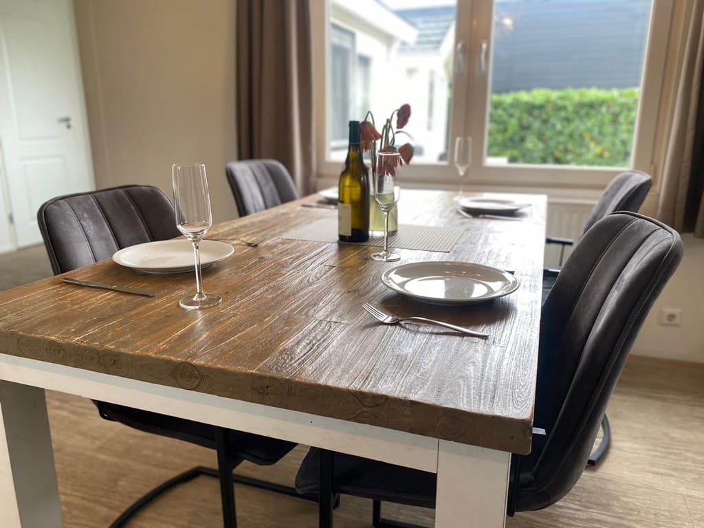 Eetkamertafel met vier stoelen, glazen, servies en flessen op een houten oppervlak bij een raam met uitzicht naar buiten