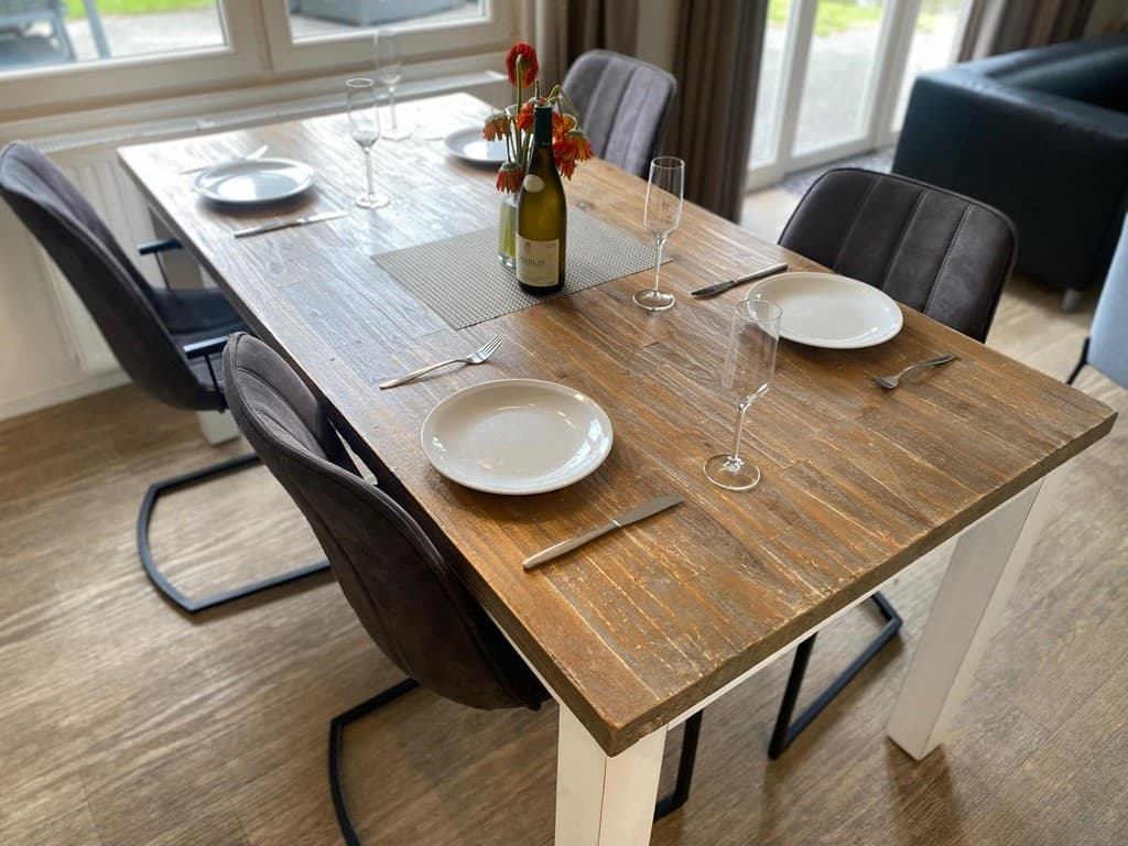Houten eettafel met stoelen, gedekte tafel met borden en glazen, bloempot met bloemen op tafel, grote ramen op achtergrond