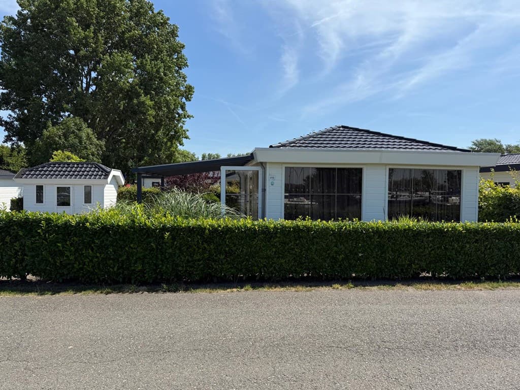Witte vakantiewoning met zwarte dakbedekking omgeven door een groene haag en bomen op de achtergrond.