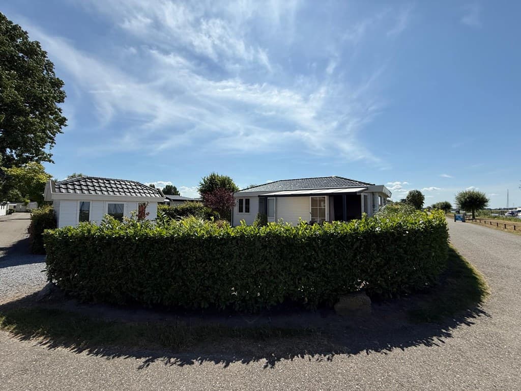 Twee vakantiewoningen omringd door groene hekken, gelegen aan een rustige straat met blauwe lucht en wolken.