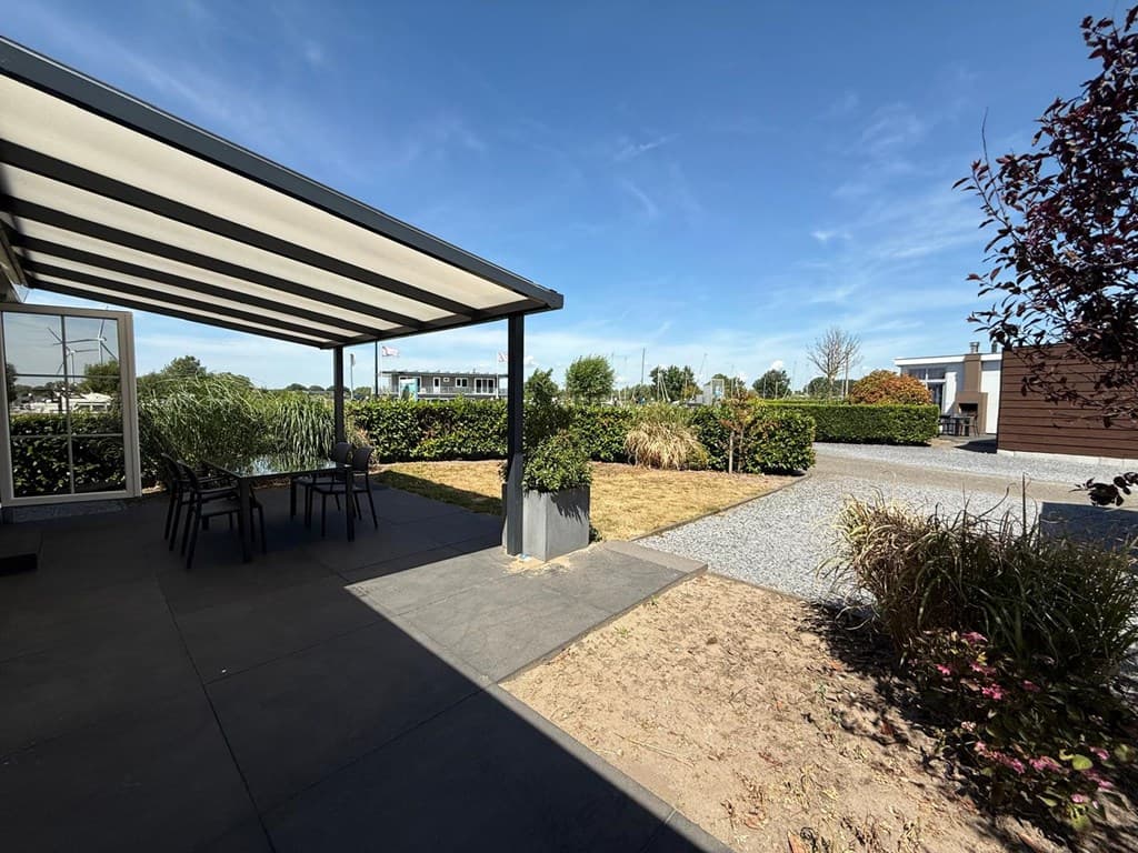 Terras met overkapping en tuin aan het water met groene planten, grindpad en zicht op omliggende woningen.