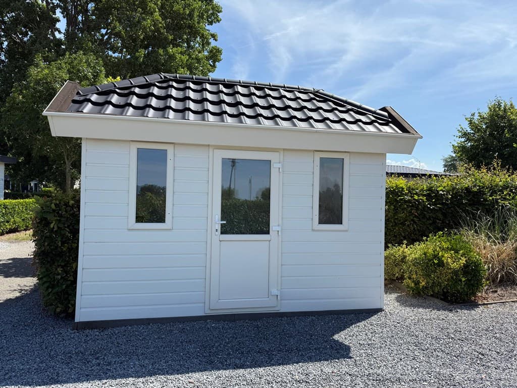 Houten vakantiewoning met een zwart pannendak en kleine ramen, omgeven door groen en grindpad.