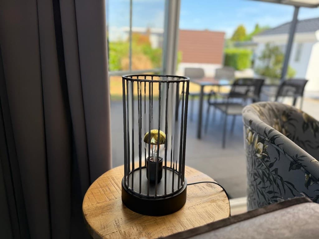 Lamp met een gouden bol in een kooi op een houten tafel, met zicht op een terras en tuin op de achtergrond