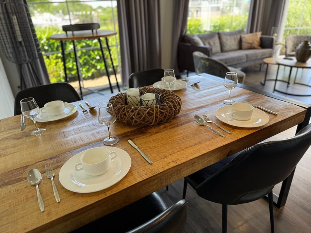 Eettafel met gedekte borden en glazen, vlakbij een bank en ramen met uitzicht op groen.