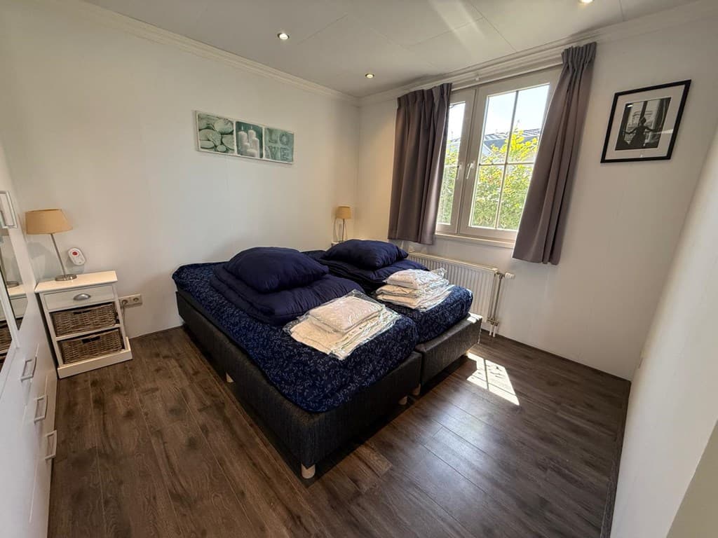 Slaapkamer met twee aparte bedden, witte muren, raam met gordijnen en een bureau met lamp en decoraties.