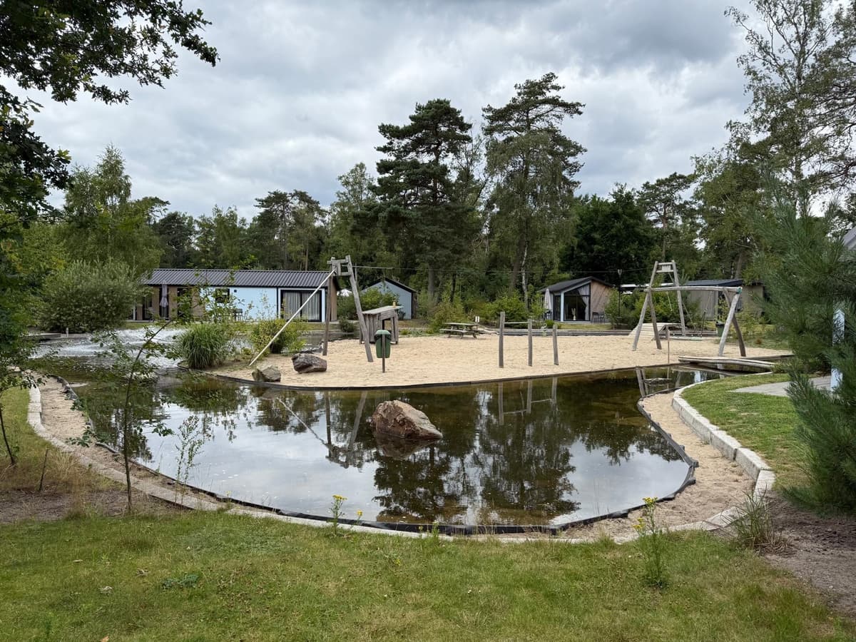 Speeltuin met zand en schommels, omringd door waterpartijen en groene bomen op EuroParcs De Hooge Veluwe.