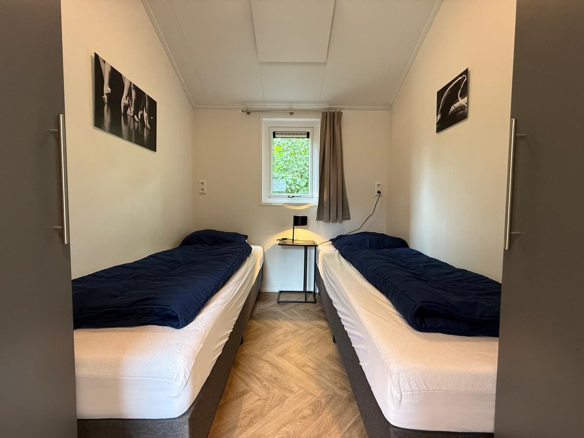 Twee eenpersoonsbedden staan in een lichte kamer met een raam en een nachtkastje tussen de bedden.