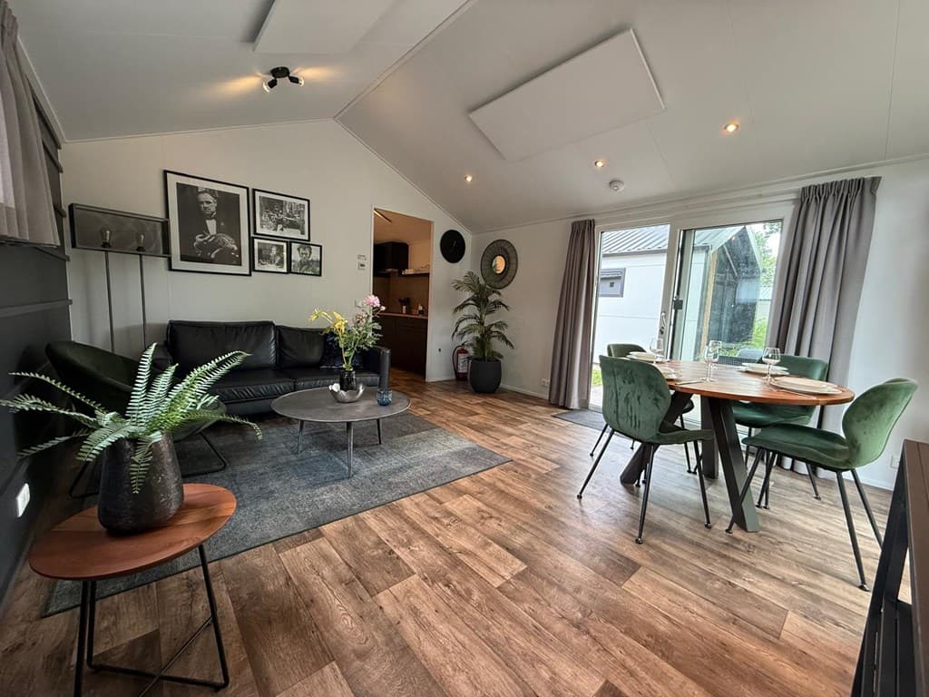 Woonkamer met zwart bankstel, eettafel met groene stoelen en grote ramen naar buiten. Decoratieve planten en kunst aan de muu