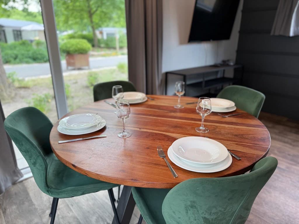 Eettafel met ronde houten blad en groene stoelen, tafel gedekt voor vier personen met borden en glazen.