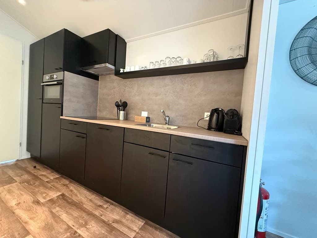 Moderne keuken met zwarte kasten, glazen en keukenapparatuur op een betonnen werkblad, witte muren en een houten vloer