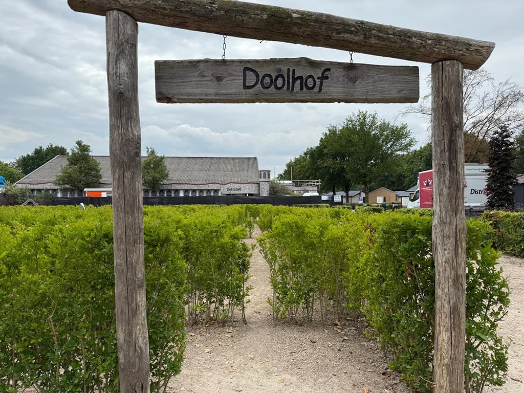 Doolhof met groene hagen en houten bord in EuroParcs Kaatsheuvel, omgeven door bomen en gebouwen op de achtergrond