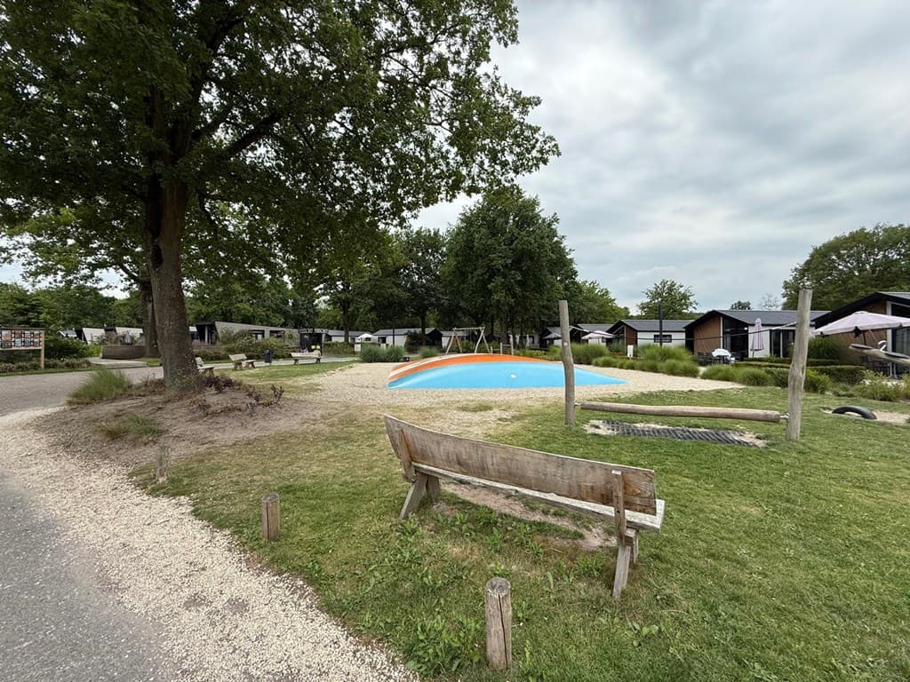 Groen veld met een speelmogelijkheid in het midden, omringd door vakantiewoningen en een bankje in de voorgrond