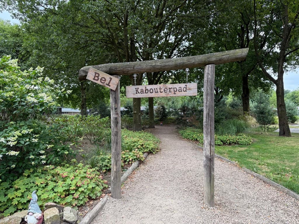 Houten bord bij een zandpad met aanwijzingen naar 'Bel' en 'Kabouterpad', omringd door groen en bomen
