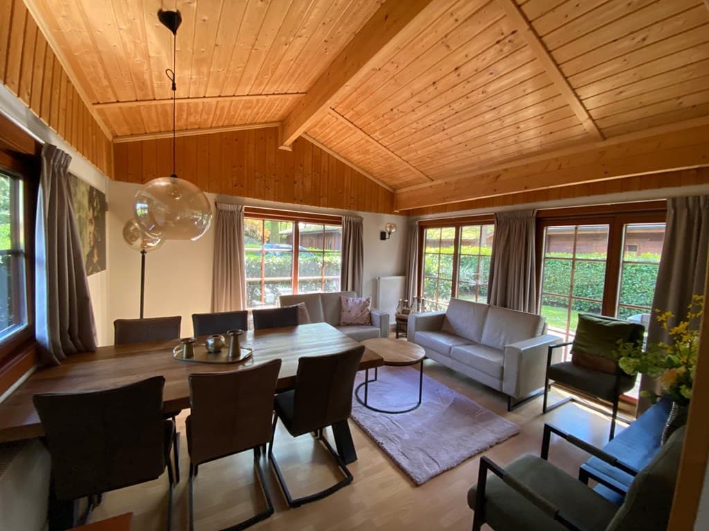 Huiselijke woonkamer met houten plafond, grote ramen en een eettafel met stoelen, naast een zithoek met banken.