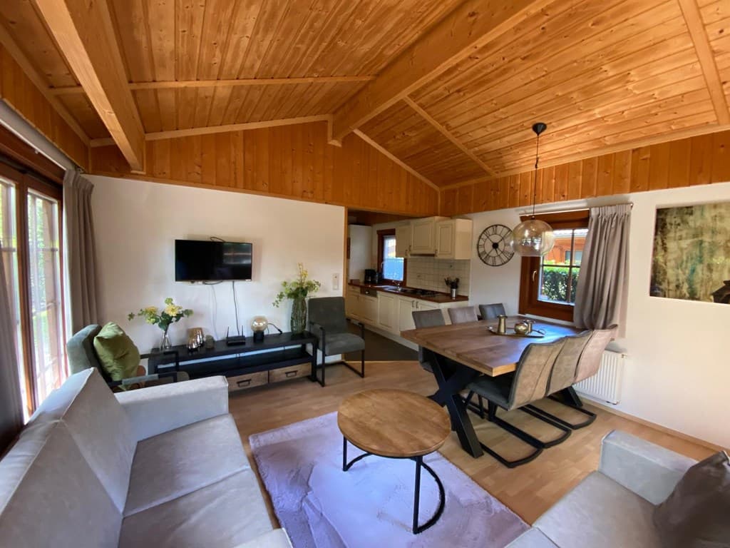 Woonkamer met houten plafond, zitbank, tv en eetkamer met grote tafel en stoelen naast de open keuken