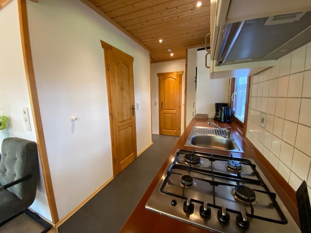 Keuken met gasfornuis, houten kasten, en links een deur naar een andere ruimte. Wand met tegelwerk en raam.