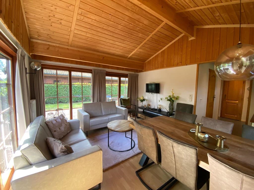 Woonkamer met houten plafond, gezellige zithoek en eettafel, grote ramen met uitzicht op een tuin.