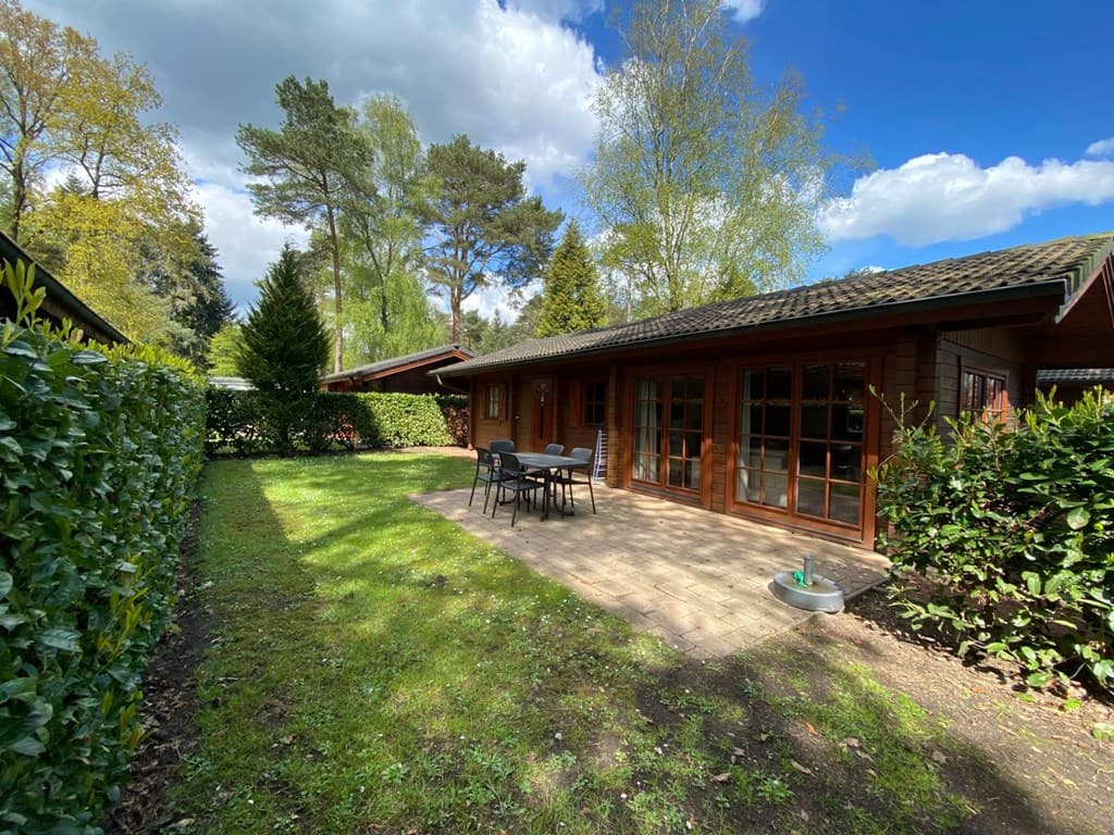 Tuin met terras bij een houten woning, omgeven door gras en struiken, met een tafel en stoelenset buiten.