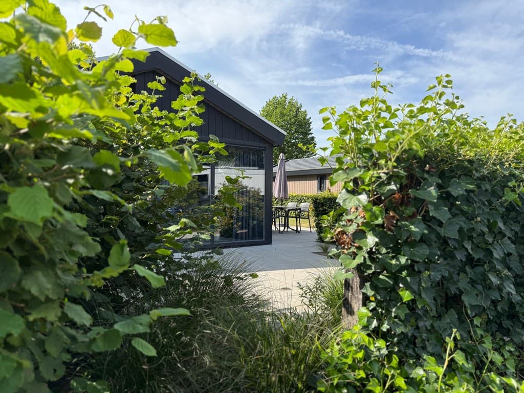 Zicht op een moderne vakantiewoning omringd door groene heggen en bomen, met een terras en parasol zichtbaar.