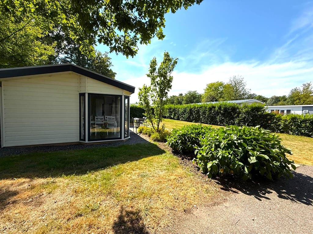 Hoek van een vakantiewoning met een groene tuin en grote ramen, omringd door struiken en bomen in een rustige omgeving
