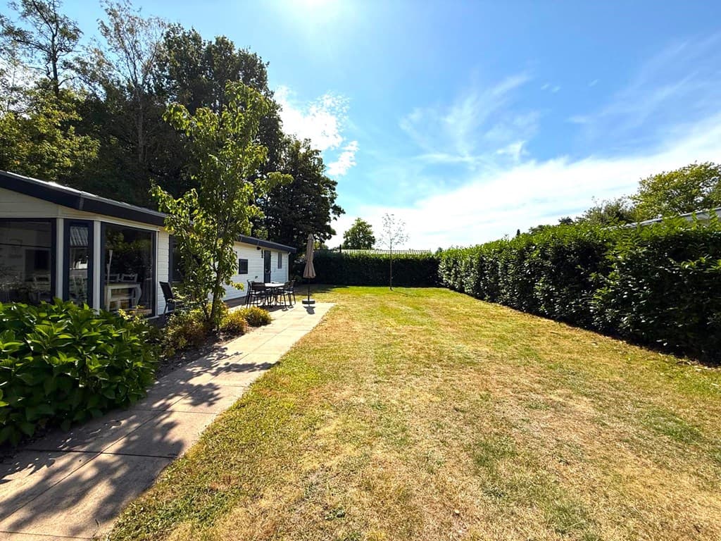 Vooraanzicht van de woning met een grasveld en border, omringd door groene hagen en bomen onder een blauwe lucht