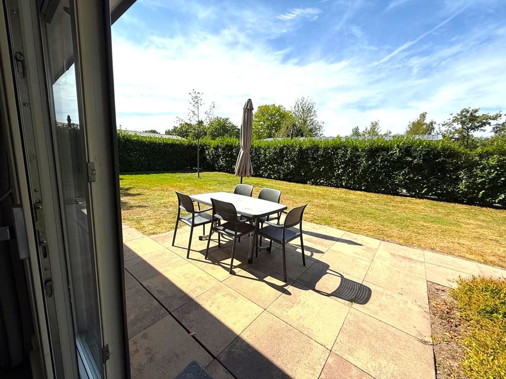 Tuin met betegeld terras, tuintafel en stoelen, omringd door gras en groene heggen.