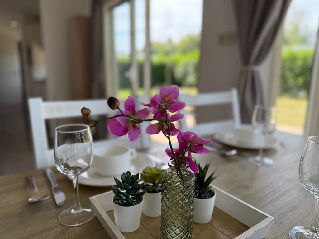 Eettafel met glazen, servies en een vaas met paarse bloemen, zicht op tuin door grote ramen.