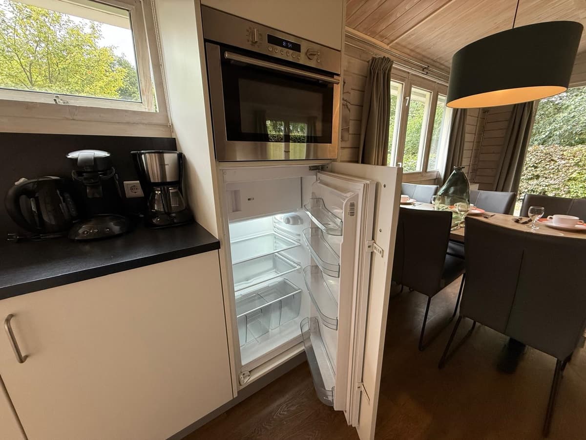 Open koelkast in een moderne keuken met koffiezetapparaat en eettafel met stoelen op de achtergrond.