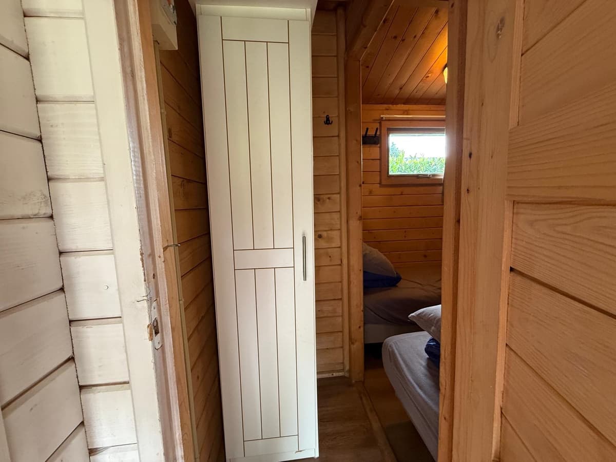 In een houten gang staat een witte kast naast een deur naar een slaapkamer met twee bedden.