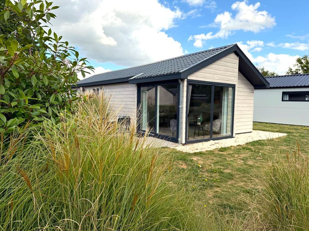 Moderne vakantiewoning met grote ramen, omringd door gras en planten, gelegen in een groene omgeving.
