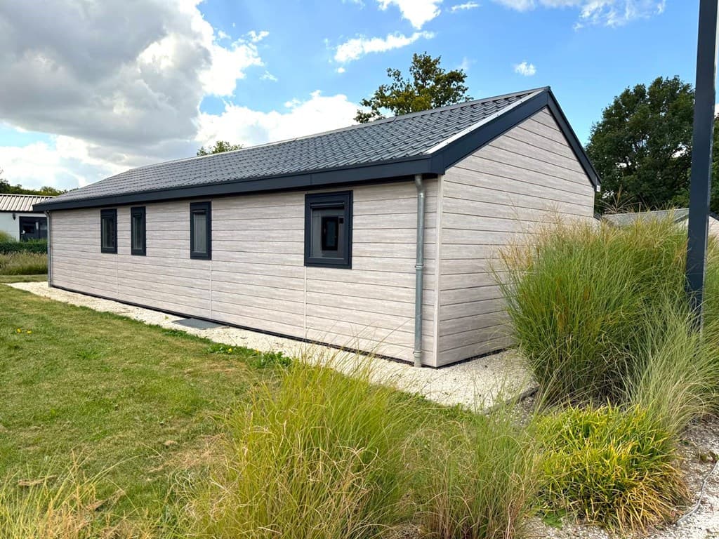Moderne vakantiewoning met lichte gevelbekleding en donkere kozijnen, omgeven door groen gras en struiken