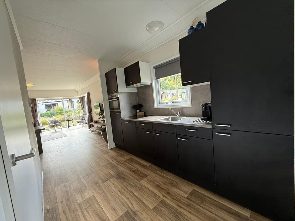 Keuken met donkere kasten, wastafel en inbouwapparaten, zicht op de woonkamer en buitenruimte met tuinmeubilair