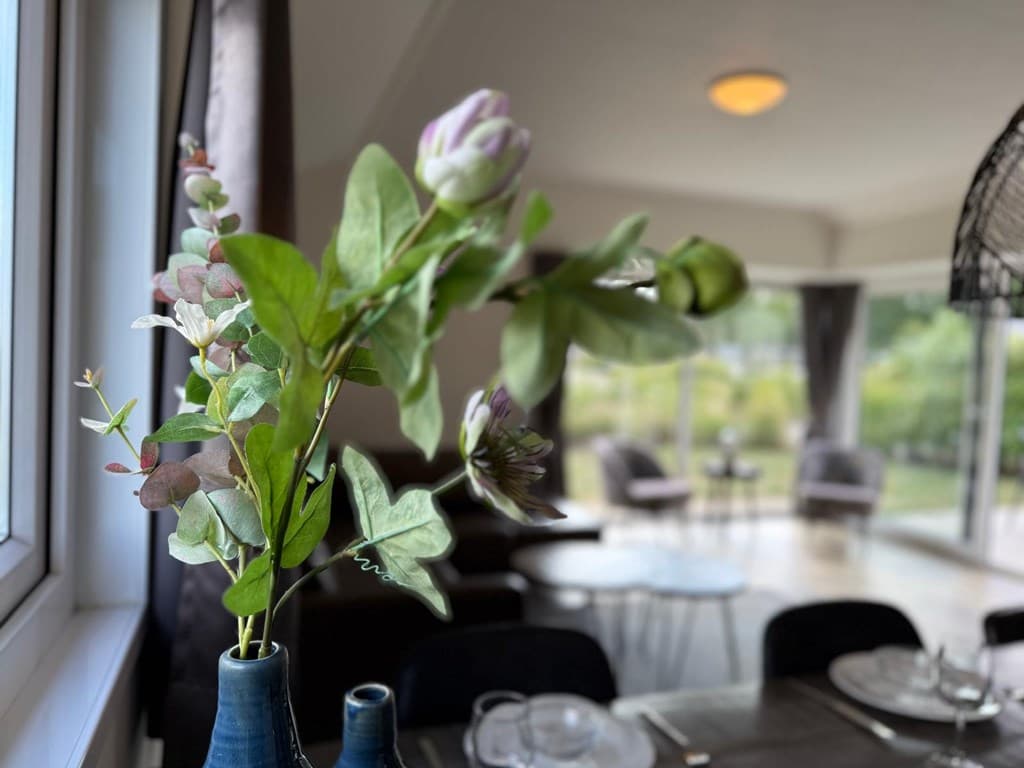 In een woning met een moderne inrichting, een vaas met groene planten en bloemen op de voorgrond en een uitzicht op de woonka