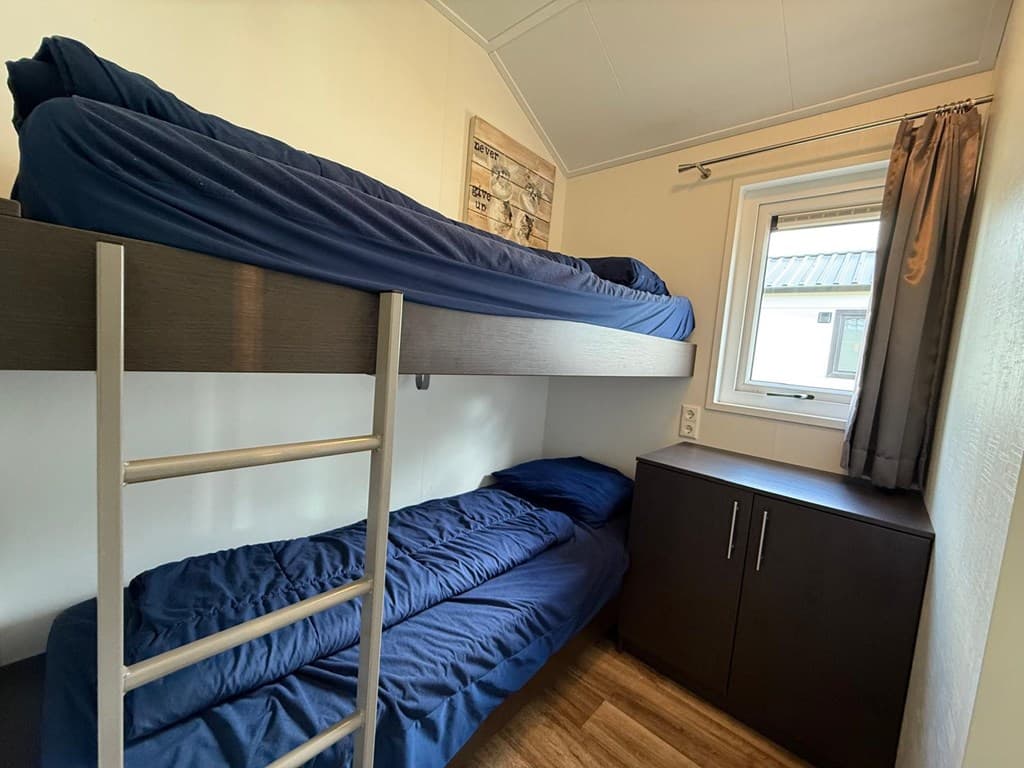 Slaapkamer met stapelbed en ladenkast, raam met gordijnen en houten vloer.
