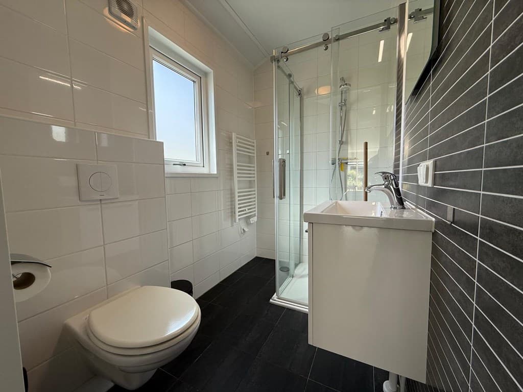 Moderne badkamer met inloopdouche, toilet, wastafel en wandverwarming in lichte tegels en donkere accentwand.