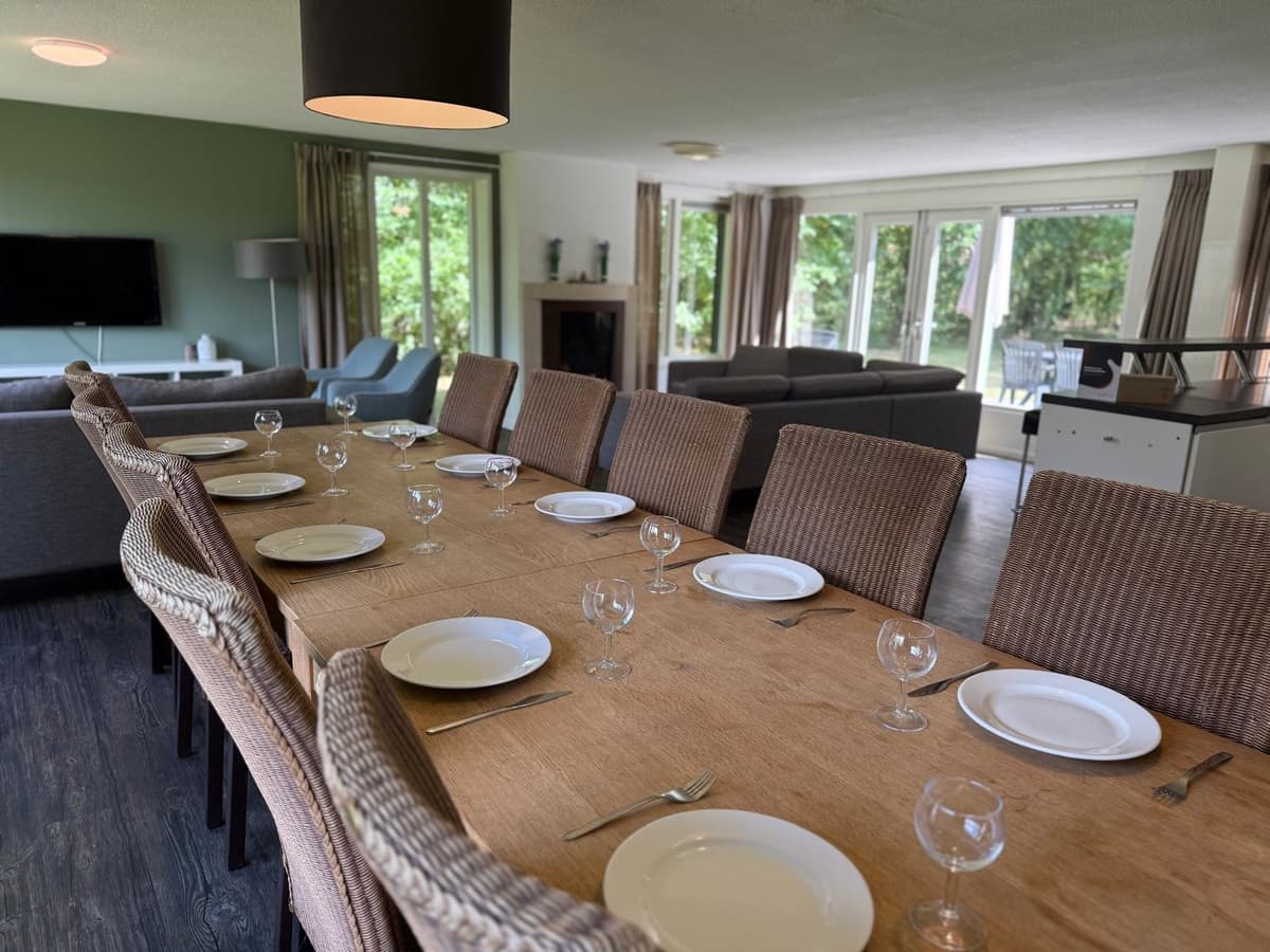 Eetkamer met lange houten tafel, stoelen en glazen, zicht op comfortabele zithoek en grote ramen naar buiten.
