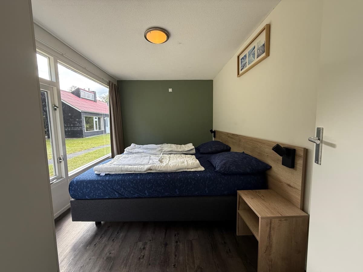 Slaapkamer met dubbel bed, nachtkastje en raam met uitzicht op een groene buitenruimte en andere woningen.