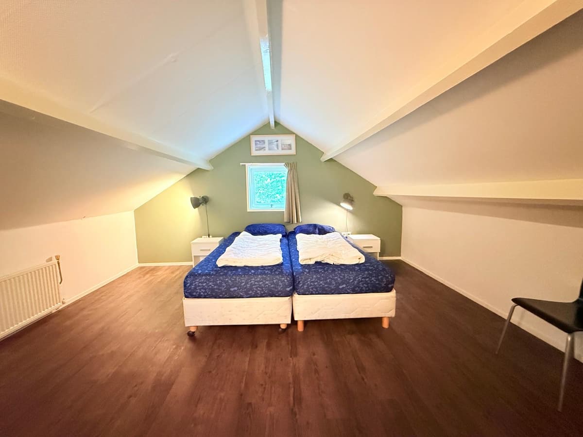 Ruimtelijke slaapkamer met twee bedden, houten vloer en schuin plafond, voorzien van een raam en nachtkastjes.