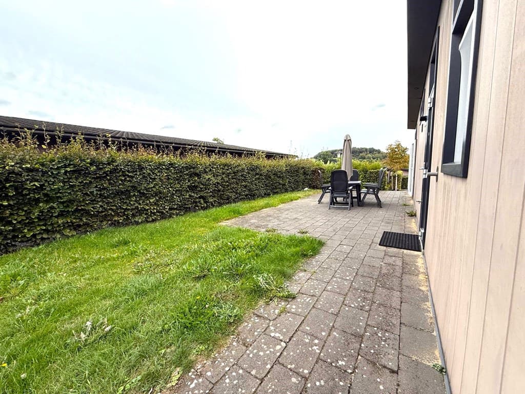 Terras met een grasveld en bestrating, omringd door groen met een parasol en tuinmeubelen zichtbaar