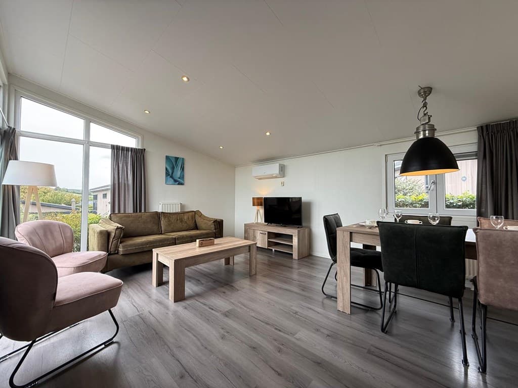 Lichte woonkamer met zithoek, eettafel en tv-unit, grote ramen met uitzicht op buitenruimte.