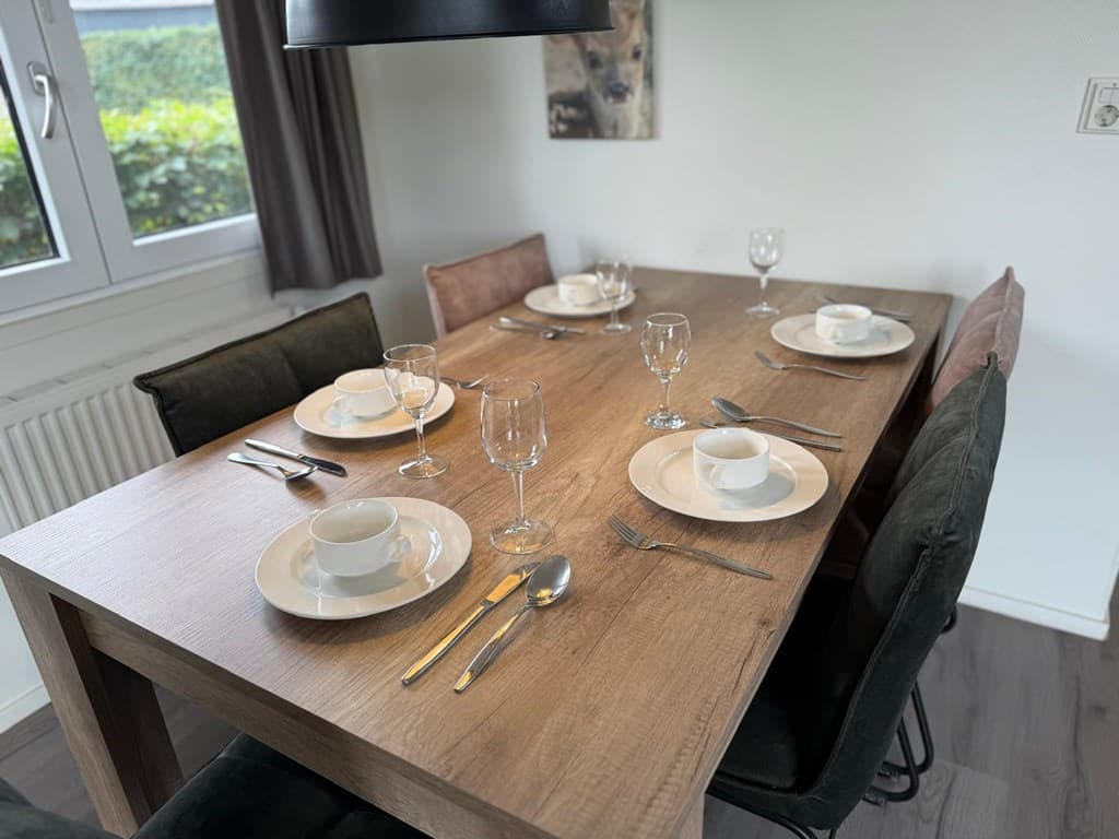 Eettafel gedekt voor vier personen met servies, glazen en bestek in een lichte ruimte met een raam en planten buiten.